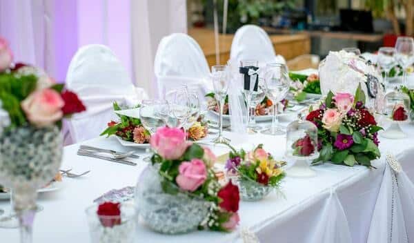 Mesa larga rectangular con mantel blanco. arriba se observan centros de mesa con rosas y flores de color rojo, rosa claro y morado. También se observan utensilios finos y copas de vidrio. Al costado de la mesa se pueden ver sillas decoradas de color blanco.