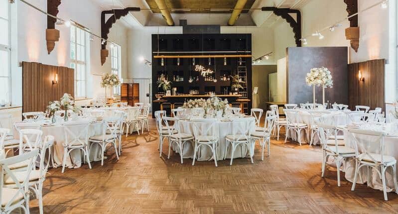 Organización mesa boda, en un salón elegante. Las cinco mesas redondas están decoradas con un mantén de color blanco, copas de vidrio y flores como centros de mesa. Las sillas son de un color blanco y se encuentran alrededor de las mesas. Detrás de las mesas se observa una pequeña cantina. Todo esta en un salón con piso de madera, elegante y llamativo.