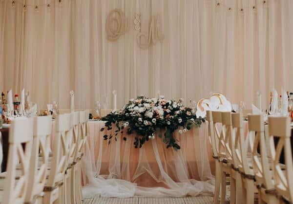 Mesa de novios es el centro decorada de colores pastel con ramo grande de flores en el centro de la mesa. A los lados hay mesas largas decoradas con utensilios finos y copas de vidrio.