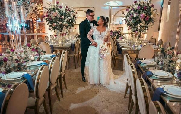 Dos novios en plena boda se encuentran de pie, mirándose de frente y el novio abrazando por detrás a la novia. A sus costados se observan mesas largas decoradas para dicho evento, con   platos de porcelana, copas de vidrio y flores de diferentes tonos de color partes en el centro de las mesas. Todo esta en un salón de un color dorado claro con buena iluminación. 