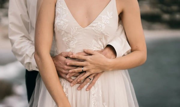 Novios en pleno matrimonio. El novio se encuentra en la parte trasera sosteniendo la cintura de la novia. La novia se encuentra por delante exponiendo su vestido de bodas sencillo pero hermoso. Ambos están de pie.