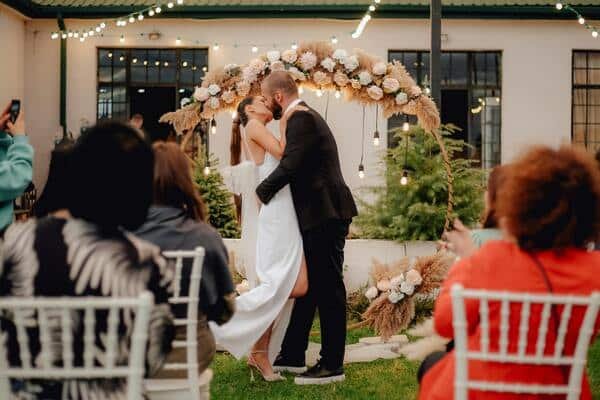 Novios besándose  en plena ceremonia de una boda. La novia viste de un vestido blanco, largo y tacones. El novio con un traje negro formal y tenis. En frente invitados sentados en sillas blancas y detrás de los novios se ve una casa decorada.