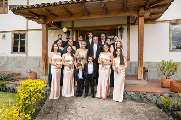 Matrimonio tomándose del brazo, rodeados de sus familiares. Las mujeres llevan vestidos color rosa claro y los hombres de traje negro elegante. De fondo terraza de casa con pequeño techo de madera.