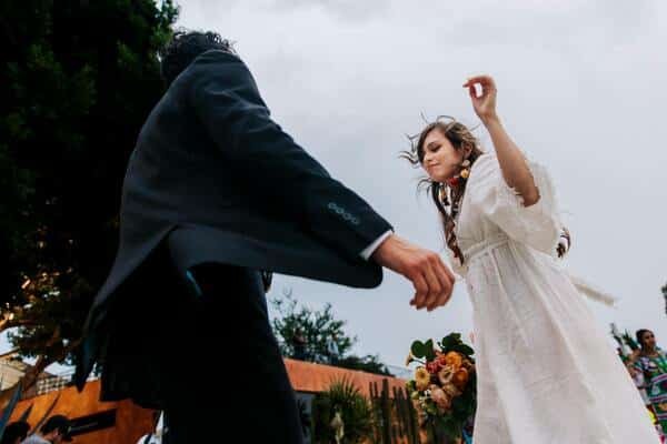 Pareja de novios bailando, la novia lleva un vestido blanco, largo y sencillos con adornos en el cabello y el novio un traje negro. De fondo una casa, árboles y cactus. 