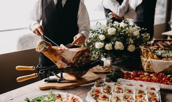 Banquete elegante con muchos aperitivos sobre la mesa. Un mesero corta con delicadeza jamón de una pierna ahumada que se encuentra sobre la mesa.