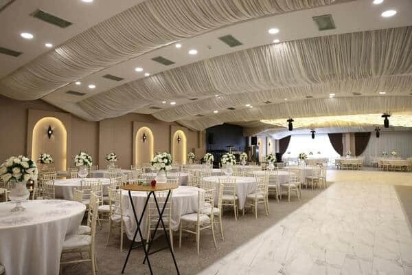 Salón decorados para celebrar un evento elegante. Arriba de las mesas centros de mesa con flores. Un pasillo largo para caminar y luces cálidas.