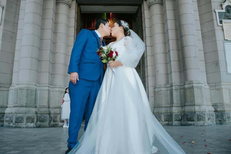 Pareja de novios besándose afuera de una iglesia grande. La novia lleva un vestido blando grande, un velo y un ramo de flores (vestido de boda). El novio un traje elegante verde con una camiseta blanca. Detrás una iglesia grande.