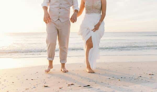 Pareja caminando por la arena del la playa. La mujer lleva un vestido largo, blanco de boda y el novio lleva un traje café claro con camiseta blanca. Los dos caminan descansos. De fondo se ve la playa, el mar y el cielo de día con pocas nubes.
