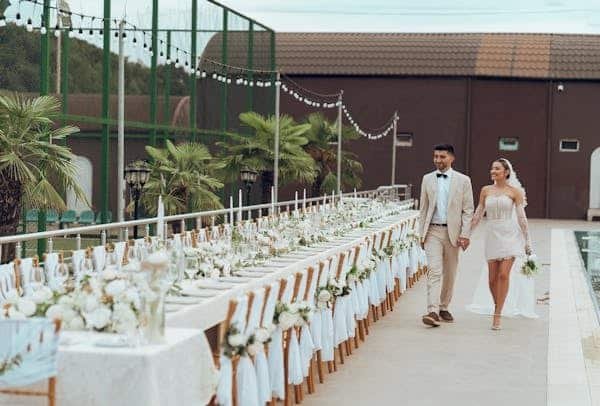 Dos novios en plena boda viendo y caminando a un lado de una mesa larga con bonita decoración de boda. La mesa tiene un mantel blanco y sobre ella se ven floreros con flores blancas, compas de vidrio y platos. Las sillas a su alrededor están decoradas con flores blancas de manera elegante. De fondo se ven palmas, luces de decoración, edificios y un campo.