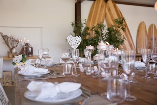 Mesa grande y rectangular de madera con vajilla de porcelana, utensilios, vasos y copas de vidrio, también se observa decoraciones de boda. De fondo una pared blanca con más decoraciones para organizar boda en casa. 