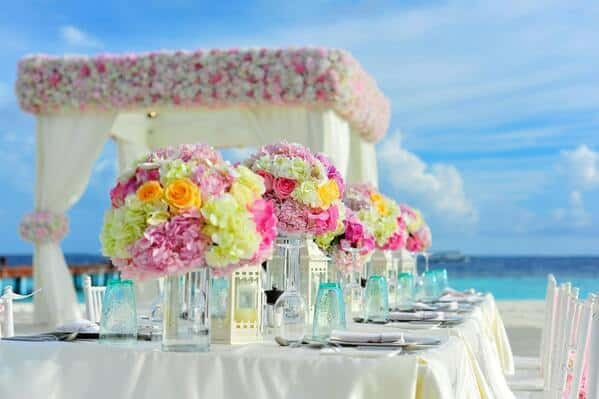 Una mesa con mantel blanco y sillas. Arriba de la mesa mucha decoración para organizar boda en la playa, floreros, vasos de vidrio y vajilla de porcelana. De fondo una carpa decorada con flores y telas. Además se ve la playa y el mar.