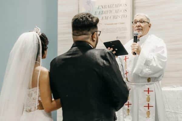 Pastor en pleno matrimonio, casando a una pareja joven en el altar. La novia lleva vestido blanco y velo, el novio un traje negro elegante, camiseta blanca y lentes. Ambos novios se ven de espalda, el pastor se ve de frente, en su mano derecha lleva la biblia y el la otra un micrófono. 