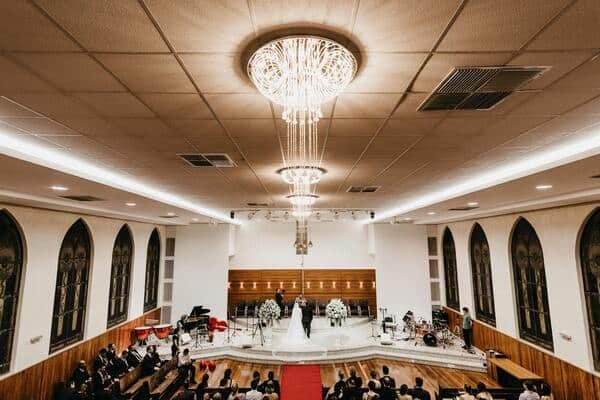 Iglesia celebrando una boda católica, se ven muchas personas sentadas y en el centro los novios. Arriba un candelabro elegante y grande. La iglesia esta bien iluminada y tiene ventanas negra grandes.