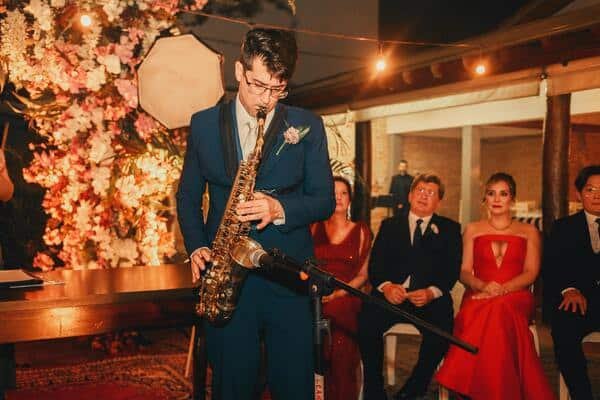 Musico tocando el saxofón en una fiesta, en medio de un grupo pequeño de personas. De fondo se ve un adorno de flores del que parece ser de boda.