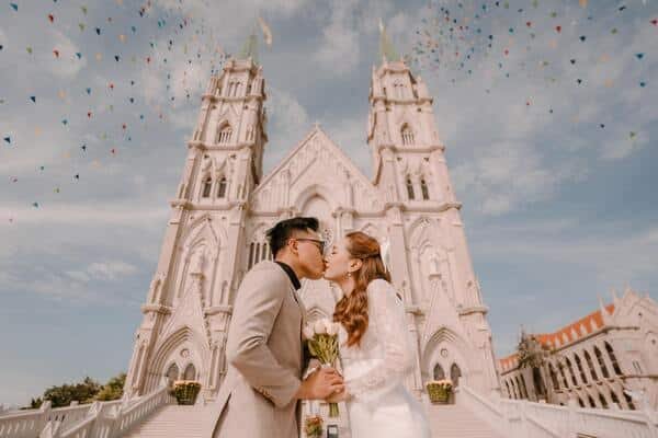 Pareja de novios besandose en frente de una iglesia grande y de color blanco. La novia lleva vestido blanco y ramo de flore, el novios un traje beige, lentes y una camiseta negra. De fondo la iglesia y banderines de colores colgados. 