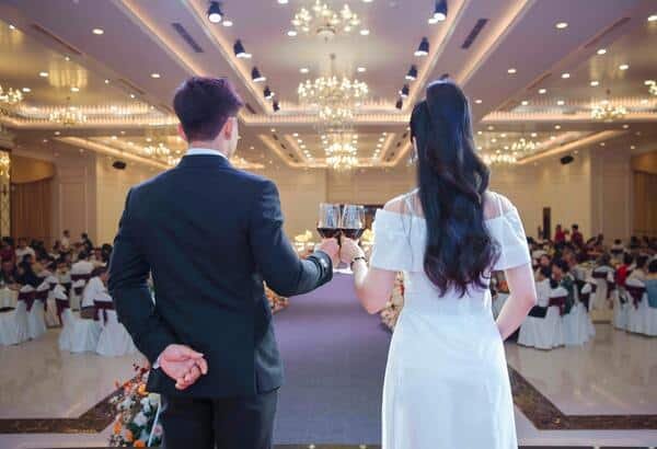 Novios en su boda, están de pie y de espalda con una copa de vino cada uno. De fondo los invitados en sus mesas observándolos.
