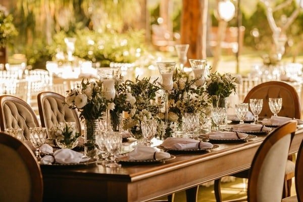 Mesa de madera organizada y decorada para una boda. Arriba de la mesa se observan copas de vidrio, vajilla de porcelana y floreros con flores blancas. Al rededor hay sillas de madera, elegantes. De fondo se ve un jardín y más mesas para invitados.