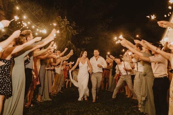 Novios caminando por en medio de varias personas que iluminan su camino con bengalas. La novia lleva un vestido blanco, largo y sencillos. El novio un pantalón café claro y camisa blanca. De fondo decoración para organizar boda en casa.