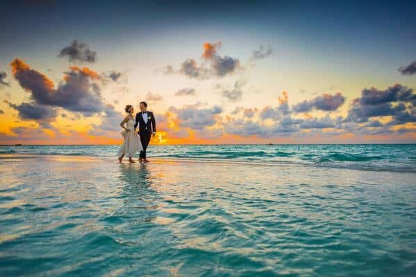 Novios caminando por la playa, tomados de la mano y mirándose con amor. Luz cálida en plena puesta de sol.
