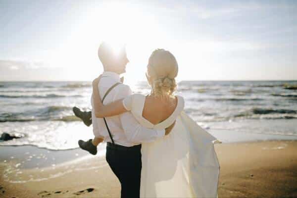 Novio carga a la novia que lleva un vestido blanco (de boda). El novio lleva una camiseta simple blanca, píntalos negro con tirantes negros. De fondo se ve la playa, el mar, arena y el sol brillante.