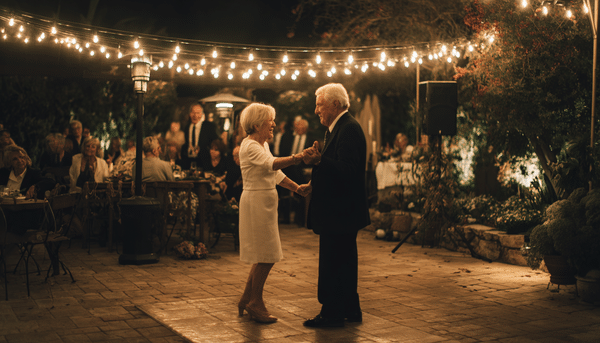 Matrimonio bailando en una pista de baile rodeados de personas. El hombre porta traje negro elegante y la mujer vestido blanco formal con tacones. La decoración es cálida con luces colgando como decoración de noche. 