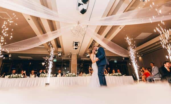 Novios bailando y besándose en medio de un salón de fiestas, celebrando su boda. Detrás de ellos se ve una mesa larga y blanca con varis invitados sentados observando a la pareja.