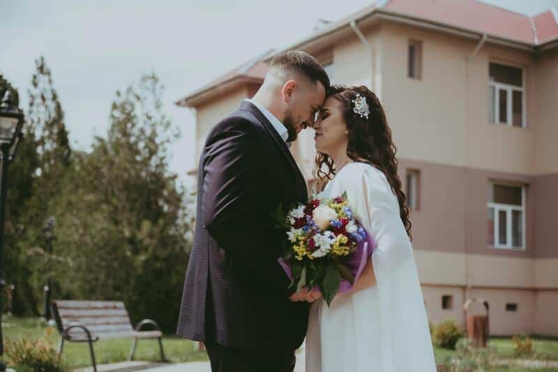 Pareja de novios de frente a punto de besarse. El novio tiene un traje negro formal y la novia un vestido blanco con un ramo de flore blancas. Detrás un casa grande, árboles, pinos y césped.