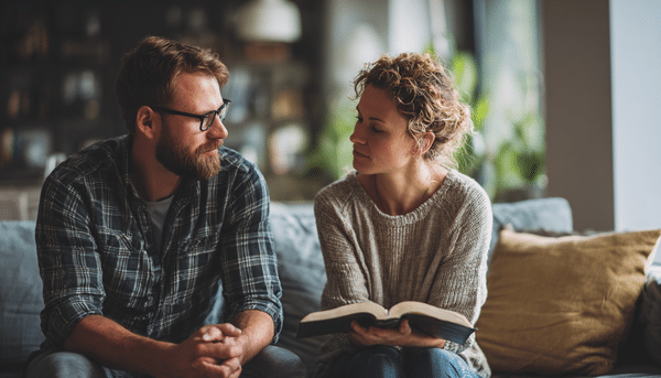 Pareja joven sentada en un sofá, orando juntos. La mujer posa en sus manos la biblia abierta y el hombre la ve fijamente. Ambos tienen un aspecto armonioso.