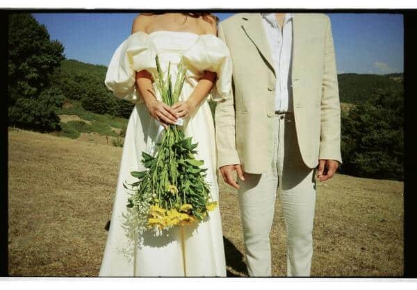 Dos novios que solo se ve su ropa. La mujer un vestido blanco cómodo, sencillo con grandes mandas y en sus manos carga un ramo de girasoles. El hombre una camiseta blanca casual y un traje café claro. De fondo se puede observar un campo.