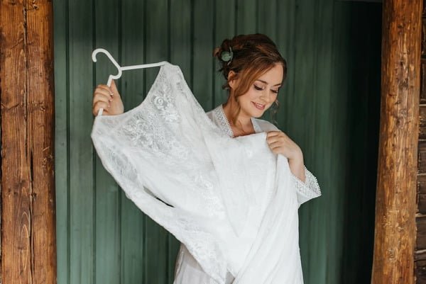 Mujer vestida con bata blanca, toma con sus manos un vestido blanco de, largo y sencillo de novia. De fondo se ve una pared de dos diferentes tomos de madera, una gris y la otra café.