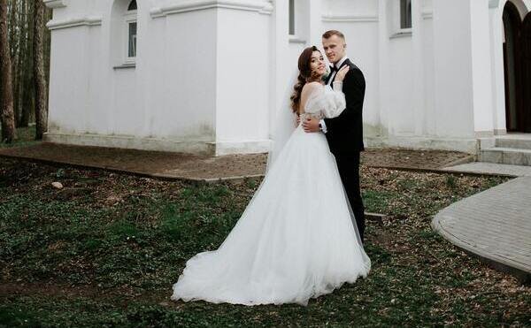 Dos novios vestidos elegantes para su boda. La novia lleva vestido blanco largo, mangas grande y un velo. El novio un traje negro, camisa blanca y moño negro. Detrás una iglesia de color blanco.